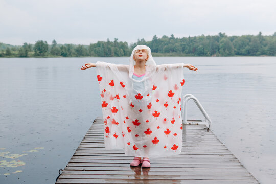 Girl child in rain poncho with red maple leaves standing on wooden lake dock. Kid rasing arms up under rain outdoors. Connection with nature. Freedom and happy childhood lifestyle.