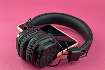 Fototapeta na wymiar Wireless black headphones and smartphone. Black over-ear headphones on pink background, soft focus