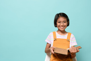 Obraz premium Asian female smile kid wear glasses looking camera and holding dictionary book while sitting chair on pastel blue background in studio. Language Learning of Schoolgirl, learning by heart concept.