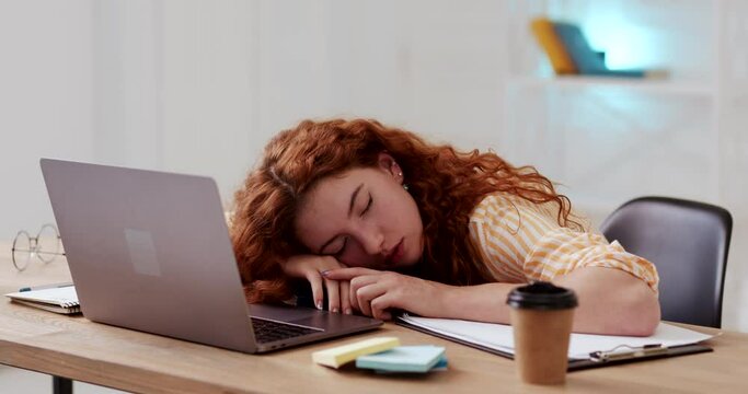 Calm relaxed young woman with red curly hair sleeping dozing peacefully on desk surface, tired of working studing on laptop, lying with head above hands.
