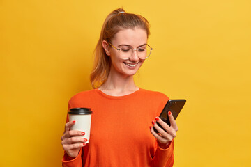 Happy pretty millennial girl downloads new mobile application, drinks coffee from paper cup, has...