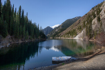 Alpine lake Kolsay in Kazakkhstan. The slopes of the mountains covered with coniferous forest are reflected in the water. A lone boat on the shore.