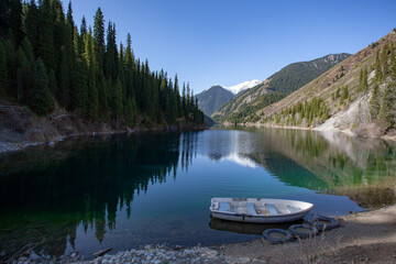 Alpine lake Kolsay in Kazakkhstan. The slopes of the mountains covered with coniferous forest are reflected in the water. A lone boat on the shore.