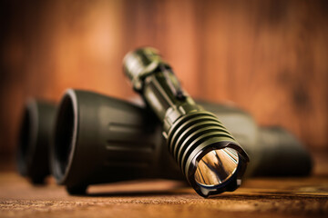 Detail close up of army - hunting binocular and flashlight on a blurred wooden background. 