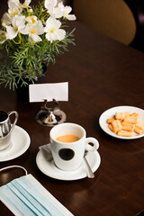 Freshly brewed hot white coffee on the wooden table inside the cafeteria with protection mask, covid-19, cookies, and metal milk jug and free space for decoration.
