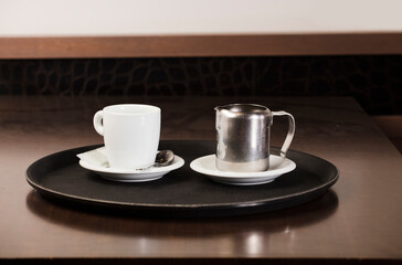 cup of freshly brewed hot coffee with metal milk container, on the waiter's tray, on the wooden table, inside the cafeteria and free space for decoration.