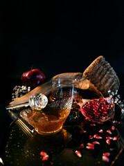 Rosh Hashanah Jewish holiday concept - blurred red apple, glass dipper on saucer of honey, Honey jar, shofar and pomegranate on black background and white flowers. Traditional holiday symbols