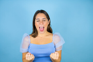 Young beautiful woman over isolated blue background very happy and excited making winner gesture with raised arms, smiling and screaming for success.