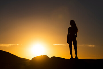 Silueta de mujer joven atractiva con pelo largo al viento al atardecer con el cielo naranja y el...