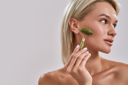 Close Up Portrait Of Young Female Blonde With Perfect Glowing Skin Looking Aside While Using Jade Facial Roller For Skincare And Beauty Treatment, Posing Isolated Over Grey Background