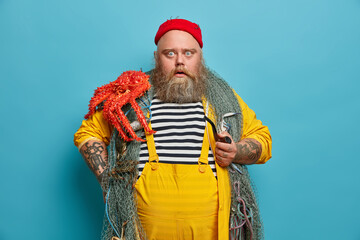 Stupefied bearded man stares bugged eyes at camera holds pipe dressed in yellow overalls red hat...