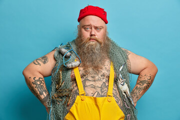 Photo of serious bearded man has confident expression keeps hands on hips, poses with fishing gears, dressed in overalls, red hat, goes agling, poses against blue background. Sea adventure concept