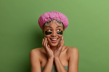 Daily skin care, wellness concept. Happy ethnic woman touches face gently, applies moisturising collagen patches after taking shower, poses bare shoulders against green background, looks aside