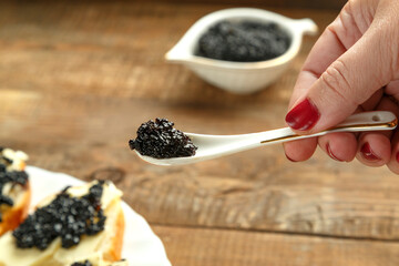 A spoon with black caviar in a woman's hand over the caviar over a white plate with sandwiches.