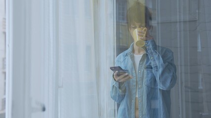 Adult woman standing in window at home daydreaming drinking coffee and checking phone at morning.