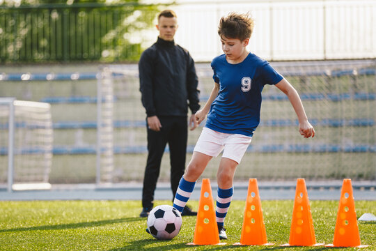 Kid soccer player dribbling through cones. Boy in soccer uniform practice with ball. Child kicking ball on grass. Young athlete improving football dribbling skills. Young soccer coach in background