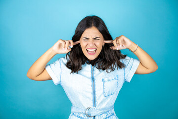 Young beautiful woman wearing a denim jumpsuit over isolated blue background covering ears with fingers with annoyed expression for the noise of loud music. Deaf concept.