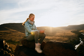 Naklejka na ściany i meble Young caucasian girl scrolling on smartphone while siting on mountain top overlooking river at sunset.