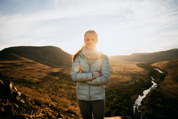 Young female standing cross armed on mountain top looking over river at susnet.