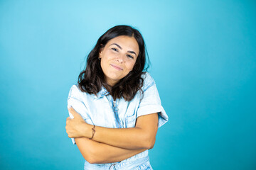 Young beautiful woman wearing a denim jumpsuit over isolated blue background hugging oneself happy and positive, smiling confident.