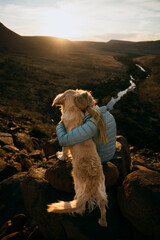 Young caucasian blonde female hugging golden dog while looking at river sitting on top of mountain.