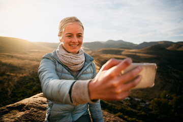 Blonde caucasian girl taking selfie on top of mountain after successful hike during sunset.