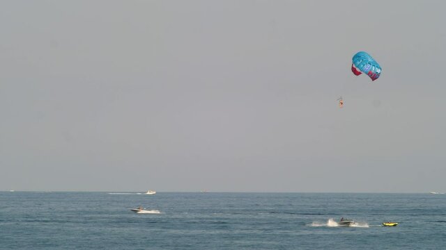 Parasailer Pulled By A Boat And Flying With A Canopy Wing Over The Sea In Nice, France. Powerboats Speeding Under The Clear Sky - wide shot