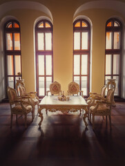 Fototapeta na wymiar Ancient royalty dining room, medieval furniture style with golden ornate chairs and table near the arched windows. Luxury hosting room background, old wood parquet. Interior architecture details.