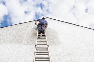 Man stands high up a ladder to paint the side of house a fresh white colour. 