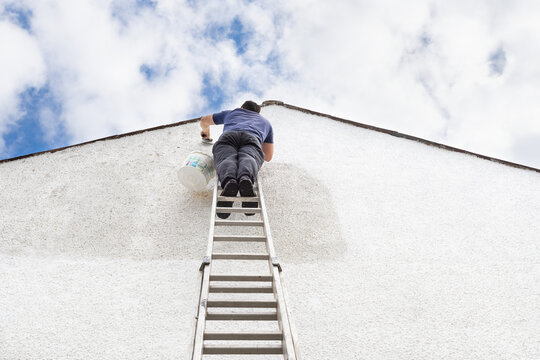 Man Stands High Up A Ladder To Paint The Side Of House A Fresh White Colour. 