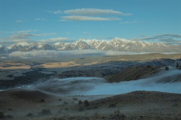 Morning landscape with a river valley, low clouds creeping on the ground and white snowy mountains in the background. Chuya ridge, Altai mountains, Siberia, Russia ..