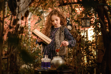 Happy Halloween Concept! Cute little witch cooking a poition in the decorated garden.  Beautiful child girl in witch costume  with halloween decoration:  book of spells, poition and big pumpkin.