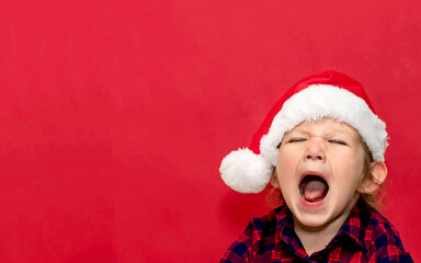 little girl yells with open mouth in red Santa hat on red background. portrait. Merry Christmas and Happy Holidays.