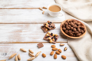 Almond in chocolate dragees in wooden plate and a cup of coffee on white wooden background. Side view, copy space.