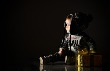 Naklejka premium Little child in gray hat, suit and boots. She looking up, holding golden gift box, sitting on floor. Black background. Close up