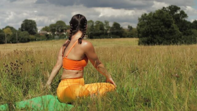 Woman sitting in long grass field doing yoga, stretching arms up to side bend to easy pose bound hands. Day time sunny UK Hertfordshire medium shot
