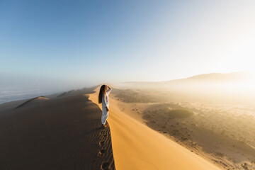 Woman enjoying sunrise on top of huge sand dune. Beautiful warm sun light and mist in morning. Sahara desert, Morocco.