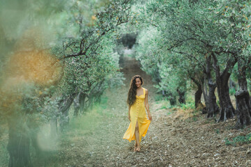Young happy smiling woman walking in olive tree garden. Yellow linen summer dress. Trendy color and textile style. - 376159313