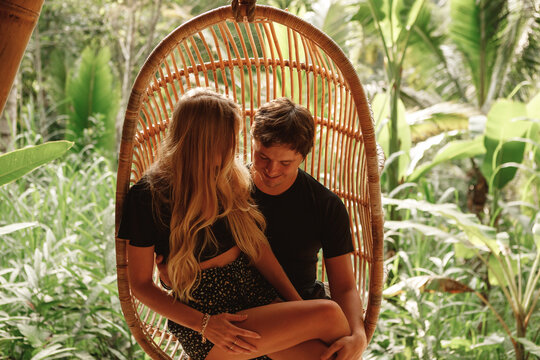 Young happy couple sit in wicker rattan hanging chair at home terrace, nature view. Romantic time