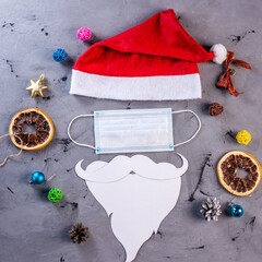 Red Santa hat, protective face mask and beard made of paper on a gray background with festive accessories. Concept of Christmas and New year during the pandemic