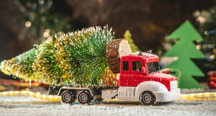 A red and white toy truck is transporting a Christmas tree against a background of lights and paper pines and snow