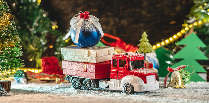 A red and white toy truck is going along a snow covered road against the background of festive lights and pines and carrying gifts. Concept of Christmas mood and preparation for the celebration