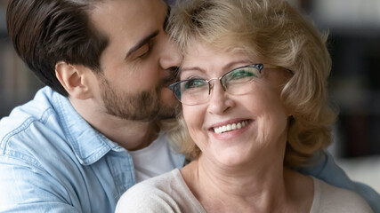 Close up loving young man kissing smiling beautiful mature mother wearing glasses, hugging from back, standing at home, overjoyed happy middle aged woman and adult son enjoying tender moment