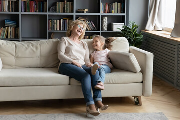 Overjoyed little granddaughter and mature grandmother wearing glasses laughing, sitting on cozy couch in living room, excited happy grandma and cute girl having fun together at home