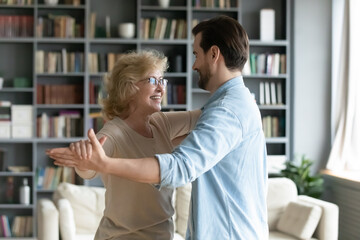Smiling mature woman wearing glasses dancing with adult son in modern living room at home, young...