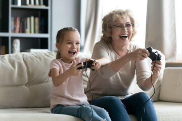 Excited laughing little granddaughter and grandmother wearing glasses playing video game together, sitting on cozy couch at home, overjoyed grandma and grandchild enjoying leisure time together