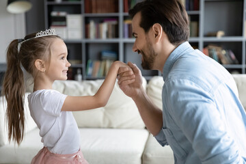 Close up loving father holding adorable little daughter hand, pretty girl wearing princess dress and diadem playing with dad, family enjoying tender moment, leisure time at home together