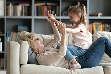 Happy mature grandmother wearing glasses playing with little granddaughter on couch, holding hands,...