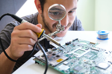 Young adult repairing a printed circuit board with a soldering iron through a magnifying glass....