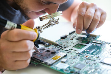 Young adult repairing a printed circuit board with a soldering iron through a magnifying glass....